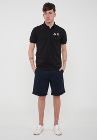 Polo nero a maniche corte, con un logo bianco, abbinato a pantaloni corti blu navy e sneakers bianche. Sfondo semplice, contesto neutro.