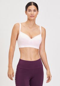 Femme debout, face à l'avant, portant un soutien-gorge de sport rose clair et un legging taille haute violet foncé sur un fond uni.