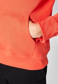 Persona vestida con un sudadera color coral, con una mano dentro del bolsillo canguro delantero, combinada con pantalones negros.