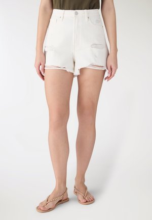 Donna che indossa shorts di denim bianchi a vita alta e strappati e sandali piatti beige con borchie, in piedi davanti a uno sfondo semplice.