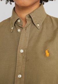 Olivově zelená košile na knoflíky vyrobená z bavlny, s bílými knoflíky a oranžovým vyšívaným logem na hrudi.