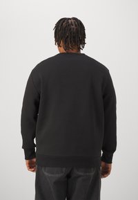 Schwarzer Pullover mit Rundhalsausschnitt, gerippten Bündchen und Saum. Glatte Textur und minimalistisches Design. Getragen mit dunkelgrauer Hose.