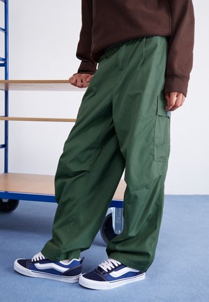Personne portant un pantalon cargo vert ample et des baskets bleu marine debout sur un tapis bleu près d'un chariot en bois.