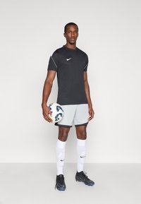 Μαύρο αθλητικό μπλουζάκι Nike, γκρι σορτς, άσπρες κάλτσες με λογότυπο, μαύρα ποδοσφαιρικά παπούτσια. Κρατάει μια μπάλα ποδοσφαίρου. Απλή φόντο.