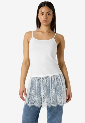 Mladá žena oblečená v bielom bezrukávovom topánku s čipkovaným lemom a modrých džínsoch, stojaca pred bielym pozadím.