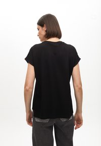 Black short-sleeve top with a curved hem and a relaxed fit. Smooth fabric with minimal stitching details. Worn with grey pants.