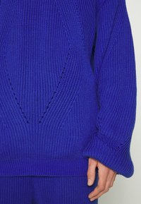 Pull-over tricoté en bleu vif, avec nervures texturées, manches surdimensionnées et découpes en forme de losange sur le devant.