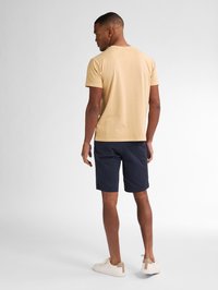 Tricou pentru bărbați cu mânecă scurtă și guler rotund, de culoare bej deschis, purtat cu pantaloni scurți bleumarin. Împerecheat cu adidași bej cu un accent maro.