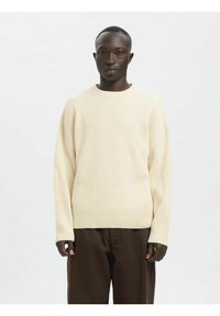 Krem pulover iz rebrastega pletenja z okroglim izrezom, spuščenimi rameni in dolgimi rokavi, kombiniran s temno rjavimi hlačami.