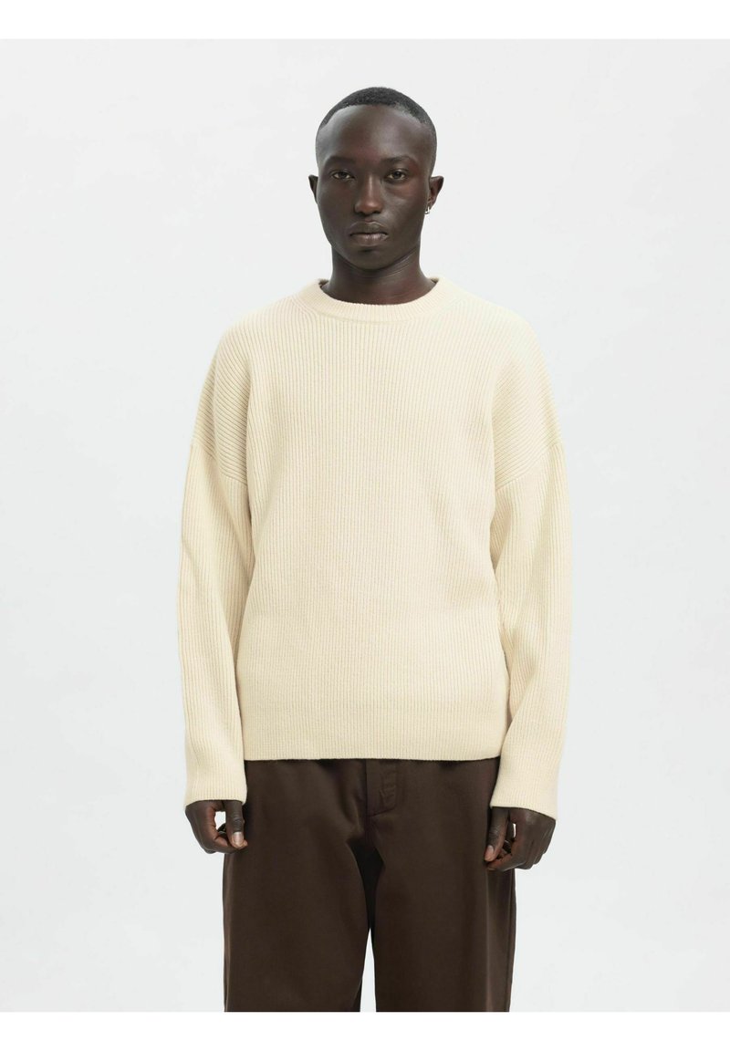 Krem pulover iz rebrastega pletenja z okroglim izrezom, spuščenimi rameni in dolgimi rokavi, kombiniran s temno rjavimi hlačami.