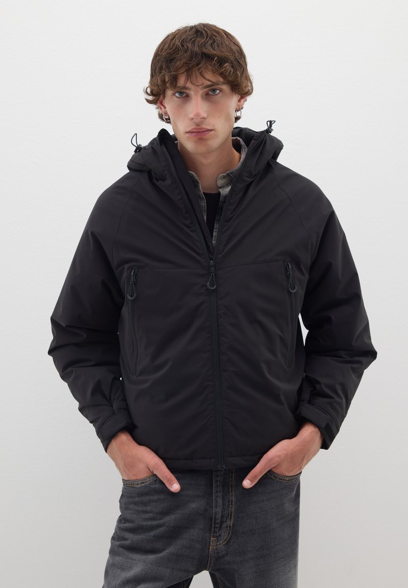 Veste noire à capuche avec poches zippées, présentant une texture lisse et des poignets élastiques, portée par-dessus une chemise en flanelle grise et un jean foncé.