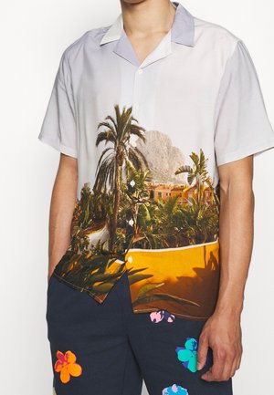 Kurzärmeliges Hemd mit Knopfleiste und tropischem Palmen- sowie Gebäudedruck, kombiniert mit dunkler Hose mit bunten Blumenmustern.