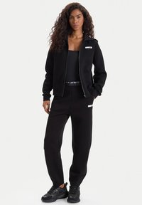Chaqueta negra con cremallera y puños acanalados, combinada con pantalones de chándal negros que tienen un bolsillo lateral y acentos de logo. Zapatillas negras completan el conjunto.