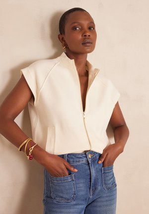 Cream, sleeveless zip-up top with a high collar, featuring side pockets and a smooth texture, paired with high-waisted blue denim jeans.