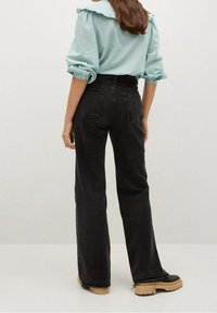 Donna che indossa una blusa azzurrina con colletto a volant, jeans neri a vita alta e a gamba larga, e scarpe platform nere con suole marroni, in piedi di spalle.