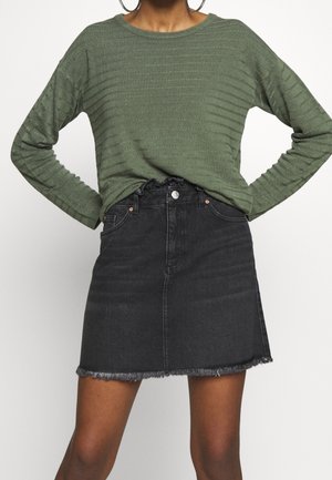 Femme portant un pull côtelé vert olive à manches longues, rentré dans une mini-jupe en denim noire taille haute avec un ourlet effiloché.