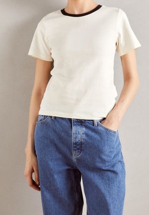 Koszulka z okrągłym dekoltem w kremowej bawełnie z ciemnobrązowym prążkowanym kołnierzem, zestawiona z niebieskimi dżinsami o luźnym kroju. Minimalistyczny design i tekstura.