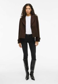 Mujer con pelo rizado que lleva una chaqueta de botones marrón oscuro, una camisa blanca, jeans ajustados negros y botas de tacón negras hasta la rodilla.