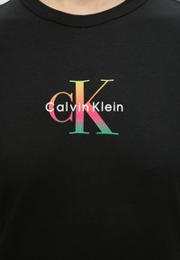 Μαύρο πουκάμισο με πολύχρωμα γράμματα "cK" σε μεγάλο μέγεθος με διαβάθμιση και λευκό κείμενο "Calvin Klein" τυπωμένο στο κέντρο του στήθους.