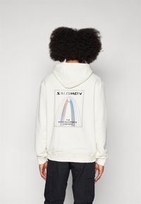 Valge kapuutsiga sviiter, millel on seljal graafika siniste, punaste ja hallide detailidega ning tekst "SALOMON THE MOUNTAIN SPORTS COMPANY."