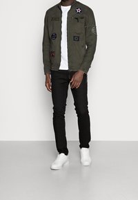 Mann in einer olivgrünen Jacke mit Patches, weißem T-Shirt, schwarzen Hosen und weißen Turnschuhen, der vor einem schlichten Hintergrund steht.