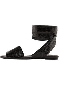 Schwarze flache Sandalen mit Krokodilprägung, offener Zehenpartie und Knöchelriemen. Sie verfügen über einen breiten Zehenriemen und ein schlankes, minimalistisches Design.
