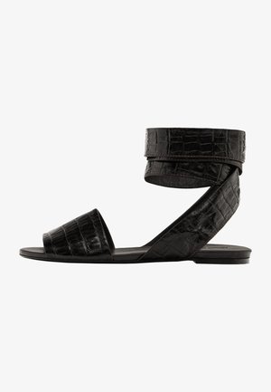 Black flat sandals with a crocodile texture, open toe, and ankle strap. Features a wide toe band and a sleek, minimalist design.