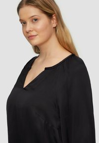 Zwarte V-hals blouse met lange mouwen en een zachte, gladde textuur. Heeft een subtiele voorkant zak en plooien bij de schouders.