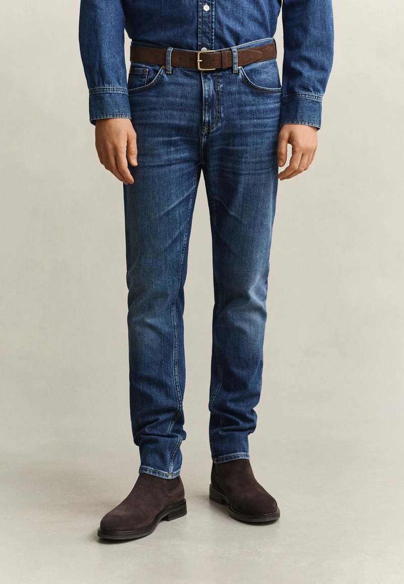 Homme portant un jean en denim bleu foncé, une ceinture en cuir marron, une chemise en denim bleu foncé assortie et des bottes en daim marron, debout sur un sol clair.