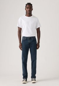 Jeans azul escuro e uma t-shirt branca. Os jeans têm um corte reto clássico, textura de ganga e design de cinco bolsos. Tênis com riscas pretas.