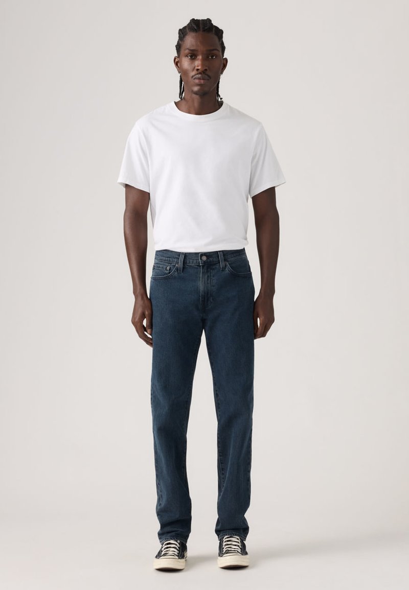 Jeans azul escuro e uma t-shirt branca. Os jeans têm um corte reto clássico, textura de ganga e design de cinco bolsos. Tênis com riscas pretas.