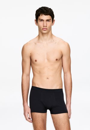 Jeune homme aux cheveux courts et foncés, debout face à l'objectif, portant un boxer noir devant un fond blanc uni.