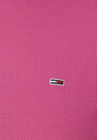 Fuchsiastrimmig tyg med en liten, broderad logotyp som har röda, vita och blå ränder på vänster bröstområde.