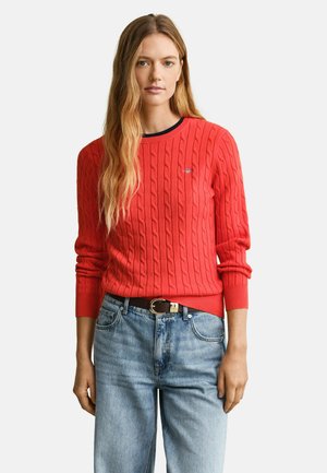 Donna con lunghi capelli biondi che indossa un maglione rosso acceso a trecce e jeans a vita alta azzurri con una cintura scura.