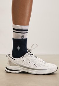 Chaussure de sport blanche avec des accents noirs, associée à des chaussettes côtelées bleu marine ornées de rayures blanches et d'un logo, sur un sol en béton lisse.