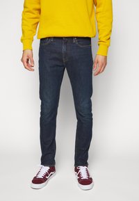 Vaqueros azul oscuro de corte ajustado, con costuras en contraste y dos bolsillos delanteros, combinados con una sudadera amarilla y zapatillas rojas.