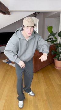 Gaiši pelēks rāvējslēdzēja džemperis ar lielām kabatām, atbilstošas pelēkas plašas bikses un sudraba sporta apavi. Bēšs cepure ar izšūtu dizainu.