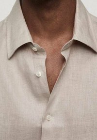 Beige linned skjorte med spids krave, frontplacket og to synlige hvide knapper, der fremviser et tekstureret stof.