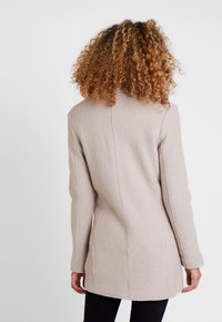 Donna con capelli ricci che indossa un cappotto di lana beige chiaro, vista da dietro su uno sfondo grigio chiaro uniforme.