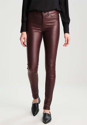 Woman wearing slim-fit burgundy leather pants, black pointed-toe shoes, and a black long-sleeve top against a plain background.