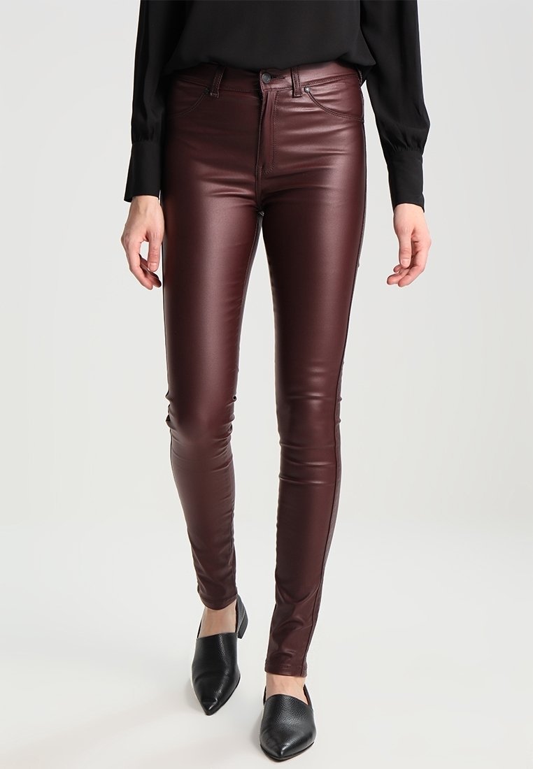 Femme portant un pantalon en cuir bordeaux ajusté, des chaussures noires à bouts pointus et un haut à manches longues noir sur un fond uni.