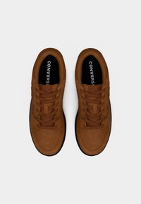 Ζευγάρι καφέ σουέντ αθλητικά παπούτσια Converse με μαύρες σόλες και μαύρα πέλματα που γράφουν "CONVERSE", φωτογραφημένα από πάνω σε λευκό φόντο.