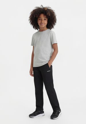 Barn med krøllet hår iført en lysegrå t-shirt, sorte bukser med logo og sorte sneakers, stående med den ene hånd i lommen.