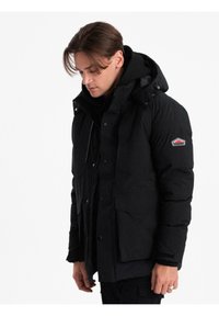 Uomo che indossa una giacca invernale nera isolata con cappuccio, tasche frontali e una toppa con il logo sulla parte superiore del braccio, con lo sguardo rivolto verso il basso.