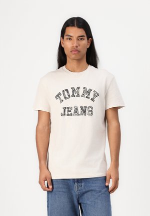 Tricou din bumbac bej cu "TOMMY JEANS" imprimat în negru pe față. Model îmbrăcat în blugi albaștri, stând pe un fundal alb.