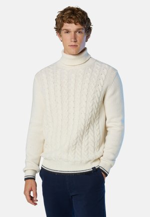 Cremfarbener Rollkragenpullover mit Zopfmuster, gerippten Bündchen und einem kontrastierenden gestreiften Saum. Getragen über einer marineblauen Hose.