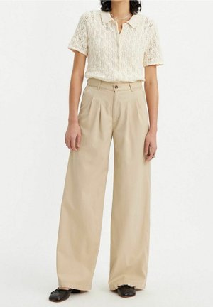 Femme portant un polo en maille crème à manches courtes, un pantalon beige taille haute à jambes larges, des ballerines noires et un collier fin en argent.