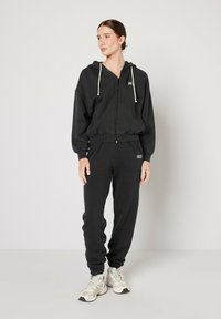 Juodas zip-up džemperis su raišteliais, derinamas su atitinkamomis sportinėmis kelnėmis. Abu pagaminti iš minkštos medžiagos, ant krūtinės ir ant kojos yra logotipas.