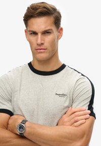 Grå bomulds-T-shirt med sort kant, korte ærmer og et lille "SuperDry" logo på venstre bryst. Modellen har en sort ur på.