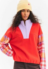 Czerwony pulower z lawendowym kołnierzem na suwak i rękawami w kratę w odcieniach pomarańczowego, różowego i żółtego. Całość uzupełnia żółta czapka z prążkowanego materiału.